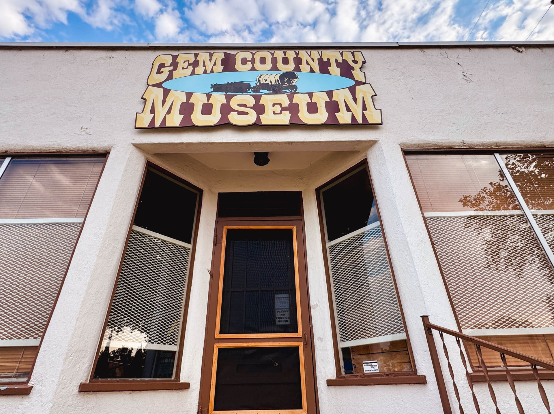 Emmett ID 3 Front entrance of the Gem County Museum with a large yellow and brown sign above the door, angled windows on either side, and a partly cloudy sky reflected in the glass. In Emmett, ID.