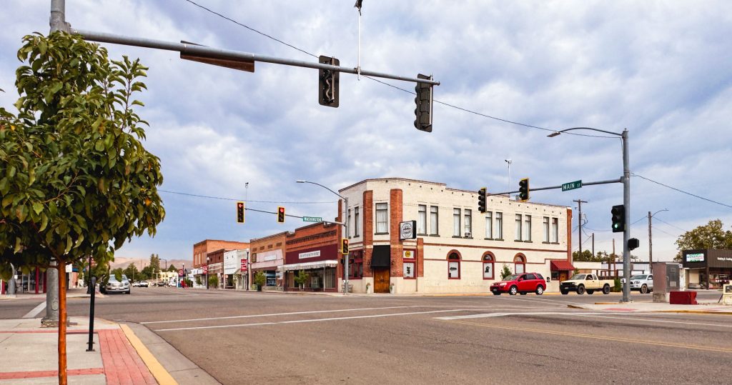 Emmett ID 26 A small town street intersection with traffic lights, a red pickup truck driving by, brick buildings with storefronts, and a tree on the sidewalk under a cloudy sky. In Emmett, ID.