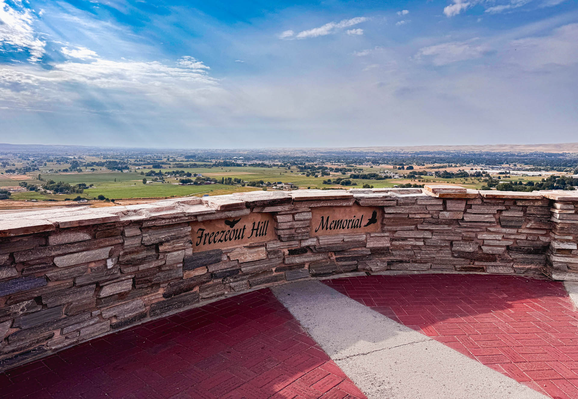 Emmett ID 5 A scenic overlook with a stone wall labeled “Freezout Hill Memorial,” offering a wide view of green fields, small buildings, and a blue sky with scattered clouds and sun rays. In Emmett, ID.