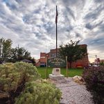 Emmett ID 23 A green park features a sign reading “Roly J. Bownan, Sr. Memorial Park” in front of a flagpole, with buildings, trees, bushes, and a dramatic cloudy sky in the background. In Emmett, ID.