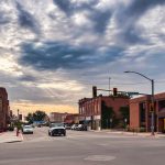 Emmett ID 24 A small-town street intersection with cars, brick buildings, and traffic lights under a partly cloudy sky at sunset. The sunlight breaks through clouds, casting a warm glow on the scene. In Emmett, ID.
