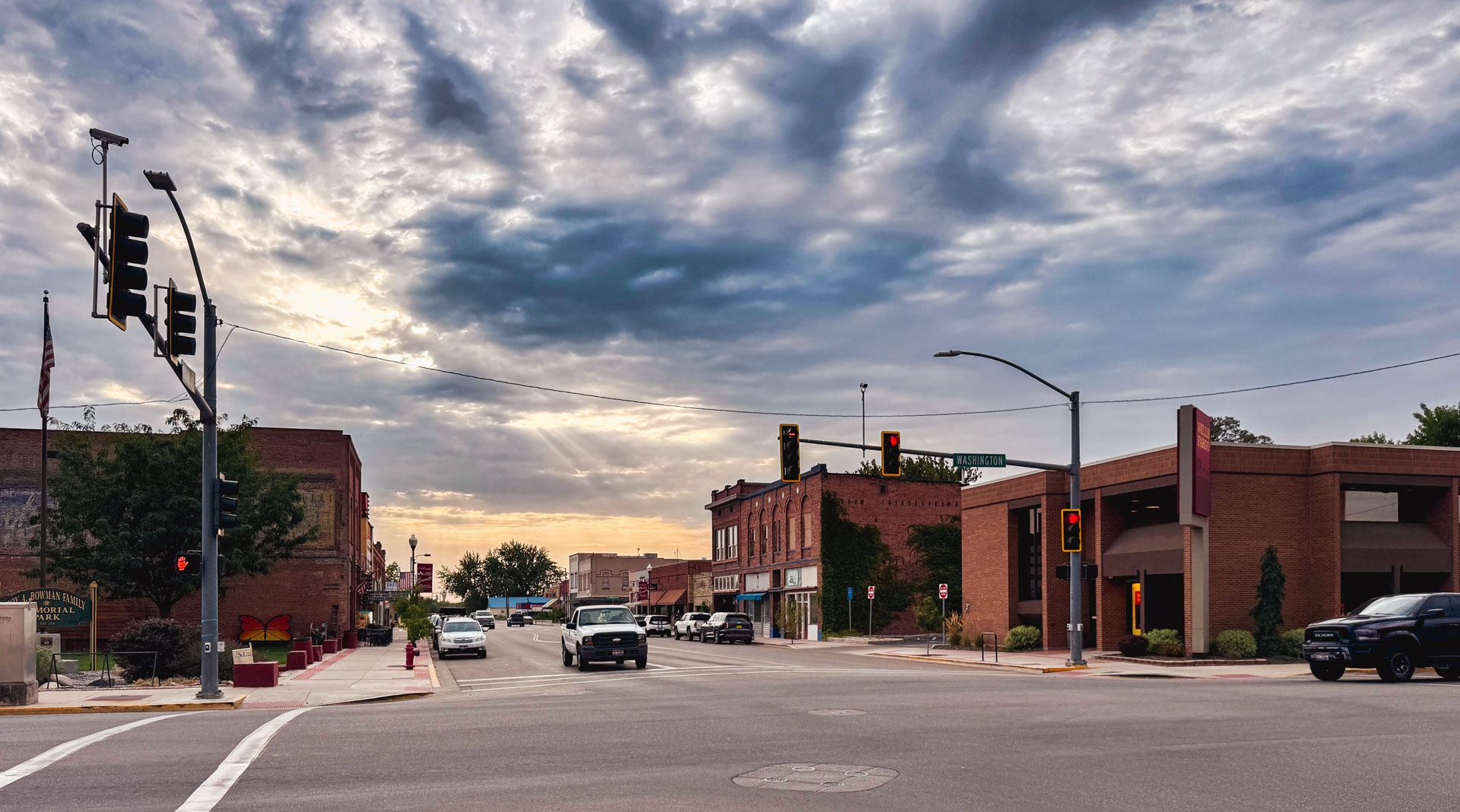 Emmett ID 4 A small-town street intersection with cars, brick buildings, and traffic lights under a partly cloudy sky at sunset. The sunlight breaks through clouds, casting a warm glow on the scene. In Emmett, ID.