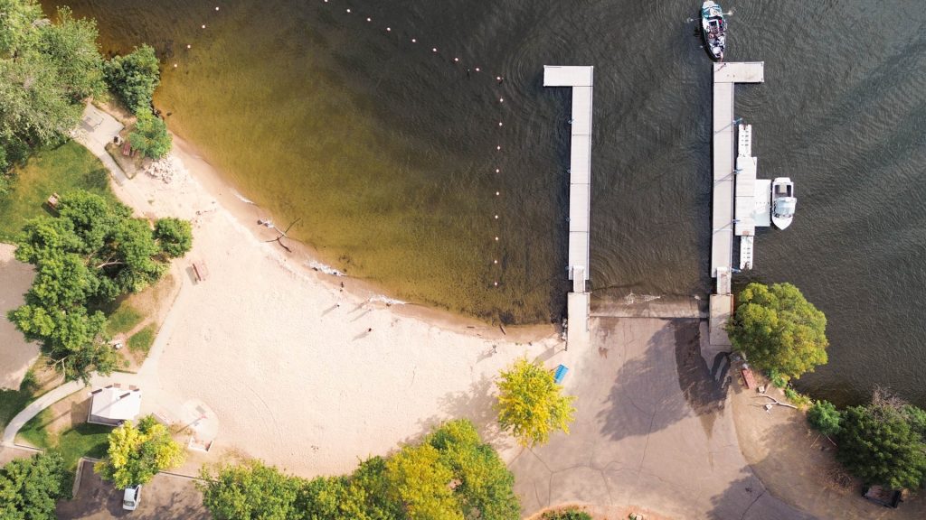 Emmett ID 6 Aerial view of a sandy beach meeting a lake, with a dock extending into the water and a boat moored at the end. Trees, pathways, and a small building surround the beach area. In Emmett, ID.