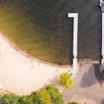 Emmett ID 10 Aerial view of a sandy beach meeting a lake, with a dock extending into the water and a boat moored at the end. Trees, pathways, and a small building surround the beach area. In Emmett, ID.