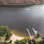 Emmett ID 13 Aerial view of a small sandy beach with a dock, surrounded by trees, next to a calm river. A boat creates a wake on the water, and hills rise in the background. In Emmett, ID.