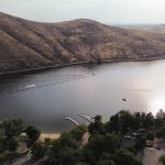 Emmett ID 11 Aerial view of a river with boats creating wake trails, surrounded by dry, grassy hills and a park area with trees, parking lots, and docks along the shoreline. The sun reflects off the water’s surface. In Emmett, ID.