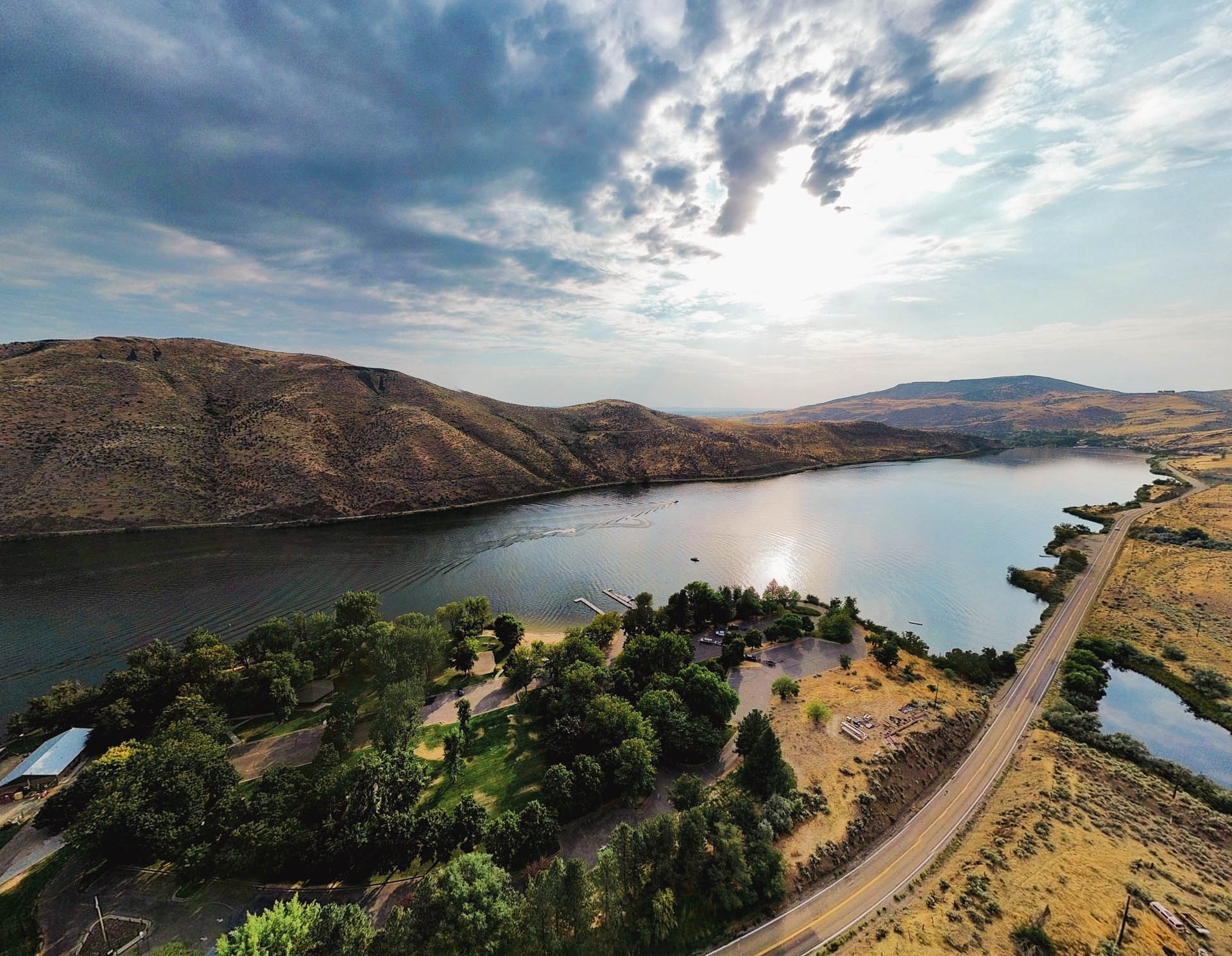 Emmett ID 2 A winding river bordered by hills and a tree-lined area with buildings, adjacent to a road curving along the water under a partly cloudy sky at sunset. In Emmett, ID.