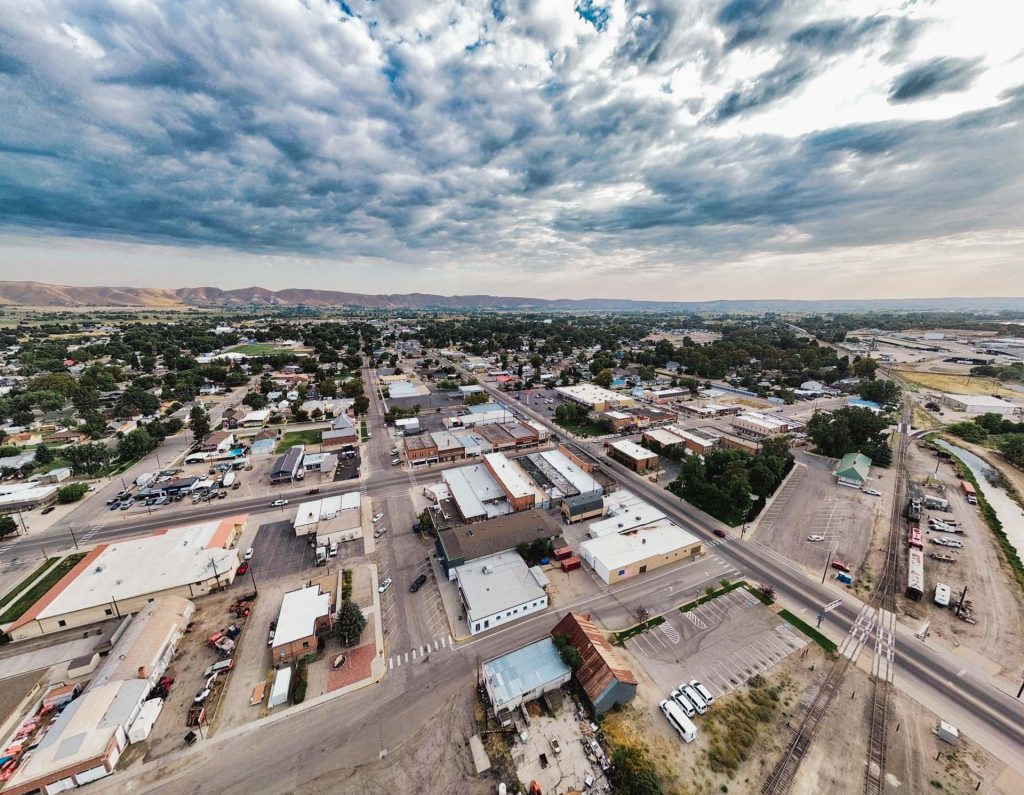 Emmett ID 15 Aerial view of a small town with grid-like streets, low-rise buildings, scattered trees, and distant hills under a cloudy sky. Roads, cars, and industrial areas are visible in the foreground. In Emmett, ID.