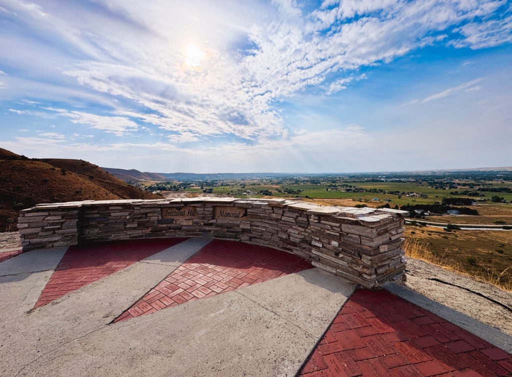 Emmett ID 25 A scenic overlook features a stone memorial wall with views of a vast green valley, distant hills, and a partly cloudy sky with the sun shining through. The foreground has red and gray patterned pavement. In Emmett, ID.