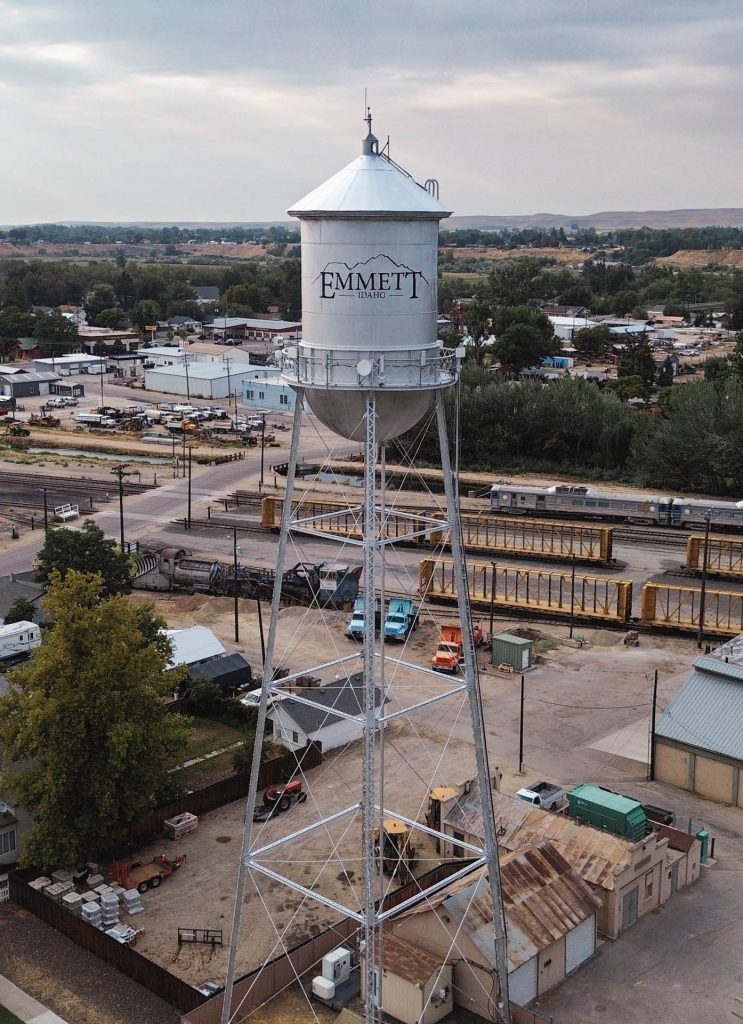 Emmett ID 1 A tall metal water tower labeled Emmett Idaho stands in a small town, surrounded by buildings, vehicles, train cars, and trees under a cloudy sky. In Emmett, ID.