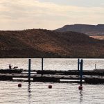 Emmett ID 12 A dock extends into a calm lake with a small boat moored and a person sitting at the end. Hills covered in dry grass rise in the background under a partially cloudy sky. Red buoys float in the water. In Emmett, ID.
